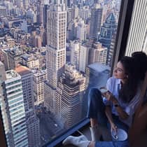 Woman with coffee cup sits in large window overlooking New York City rooftops