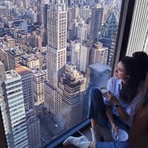 Woman with coffee cup sits in large window overlooking New York City rooftops