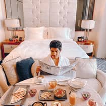 Man wearing bath robe reads newspaper in front of breakfast table, hotel bed