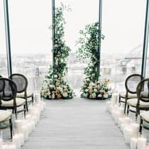 An indoor wedding ceremony set-up with tall floral arrangements, candles lining the aisle and a large floor-to-ceiling window overlooking a city skyline