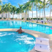 Man in spa pool looks out over outdoor swimming pool, row of palm trees