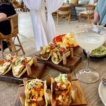 A dining table on a patio filled with various tacos served on wooden boards, fresh guacamole and a chilled cocktail