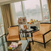 A woman dressed a white bathrobe reads a newspaper while seated at a dining table filled with breakfast food overlooking a harbor view
