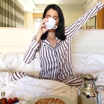 Woman on bed sips coffee cup, stretches in front of in-room breakfast dining tray