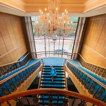 Girl in blue dress on grand blue staircase with chandelier overhead.