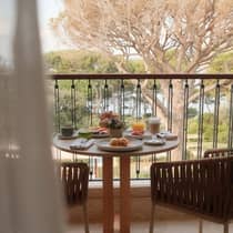 Breakfast dishes such as pastries, fruit, coffee and juice sit atop a table on a balcony overlooking lush greenery and water