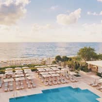 Aerial view of an outdoor swimming pool, rows of chairs, and umbrellas on the deck near the beach and ocean, creating a picturesque seaside escape.