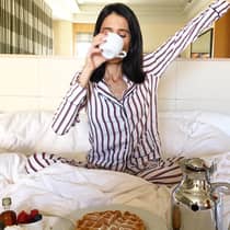 Woman on bed sips coffee cup, stretches in front of in-room breakfast dining tray