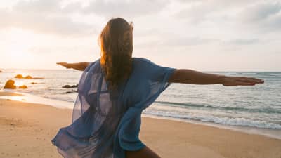 Woman wearing sheer beach cover kneels with arms outstretched in yoga pose on sandy beach by water