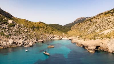 Aerial view of two boats in variegated azure water under a clear blue sky in a cove framed by rocky hills dotted with shrubs.