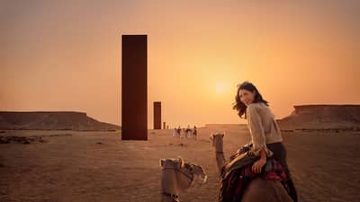 Person riding a camel looks over their shoulder and smiles with a row of black monoliths rising out of the desert landscape in front of them, all lit by an orange glow from the sunset
