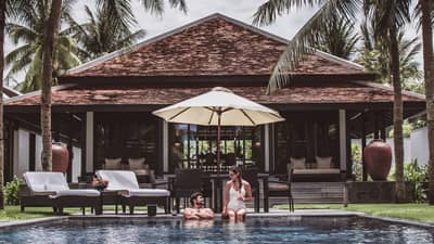 Man, woman in swimsuits at edge of outdoor pool under patio umbrella, by villa