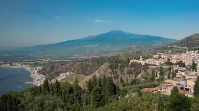 Panoramic view of a coastal town nestled in the hills, overlooking the sea, with mountains in the background and greenery in the foreground.