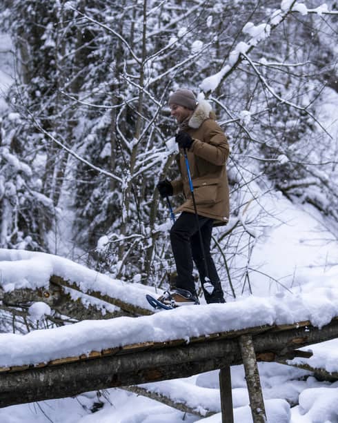 A snowshoer in a knit cap, parka and gloves uses poles to cross fallen logs in a dense, snow-covered forest.