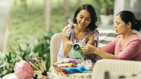 Two people engaged in a traditional craft activity, working together on fabric art while seated on a wicker patio set in a lush, green outdoor setting.