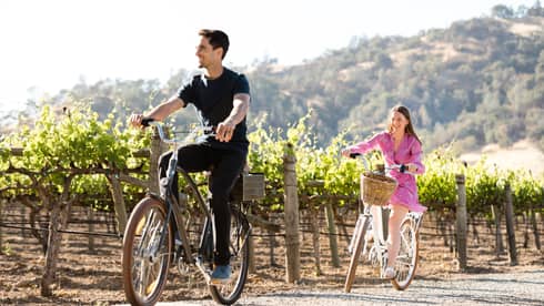 A couple rides bicycles down a dirt path flanked by beautiful vineyards, enjoying the sunshine outdoors.