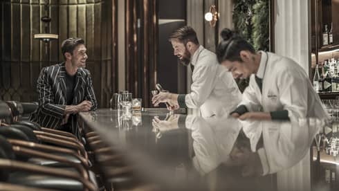 Bartender preparing a drink while a customer sits at the bar, both smiling, in a modern, upscale bar with a reflective countertop and warm lighting.