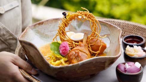 Bowl of curry noodle soup with chicken drumsticks, crispy noodles, boiled egg and garnish, served with condiments on the side.