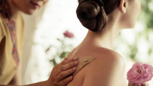 Spa staff rubs thick lotion on woman's bare shoulders as she holds a fresh flower