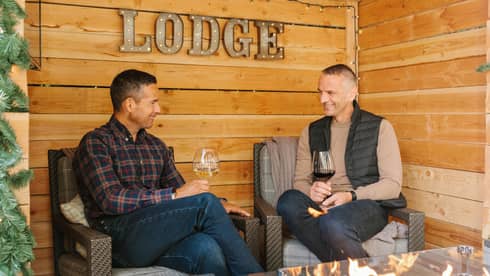 Two men drinking wine in a small cabin-like backdrop.
