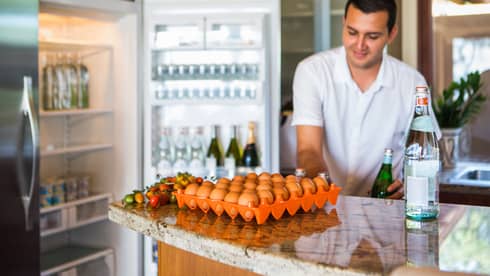 Smiling man holds green bottle in front of open fridge, fresh eggs on counter