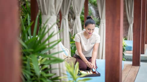 Spa attendant displays small bottle of oil, holds tray over patio lounge chair on sunny day