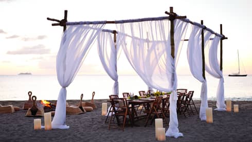 Sheer white curtains draped across wood posts around dining table on beach at sunset