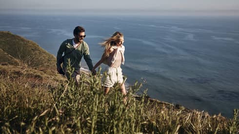 The sun shining on a smiling couple holding hands as they hike along a grassy cliff. Open water ripples below them.