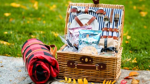 A picnic basket with plates, utensils and snacks resting on a stone near grass and yellow leaves.