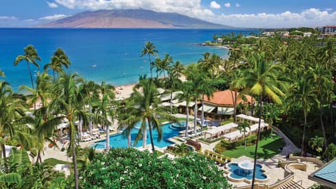Aerial view of resort pool, patios and palm trees on ocean, mountain in background