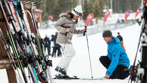 Hotel staff holds skis as woman holding poles steps in by ski rack, snowy hill