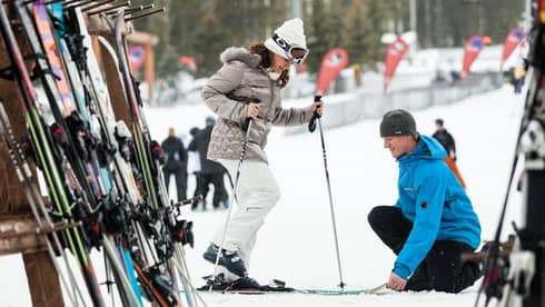 Hotel staff holds skis as woman holding poles steps in by ski rack, snowy hill