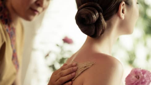 Spa staff rubs thick lotion on woman's bare shoulders as she holds a fresh flower