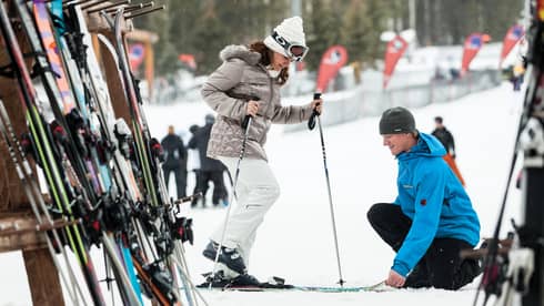Hotel staff holds skis as woman holding poles steps in by ski rack, snowy hill