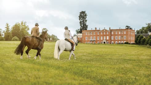 Couple rides horses across green English field to Four Seasons Hotel Hampshire building