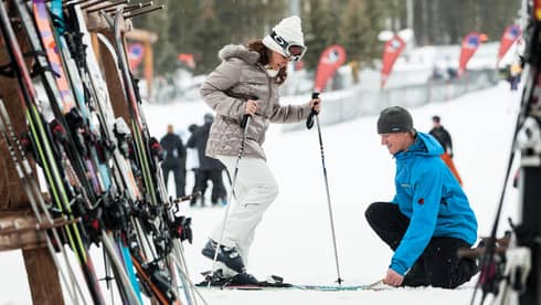 Hotel staff holds skis as woman holding poles steps in by ski rack, snowy hill