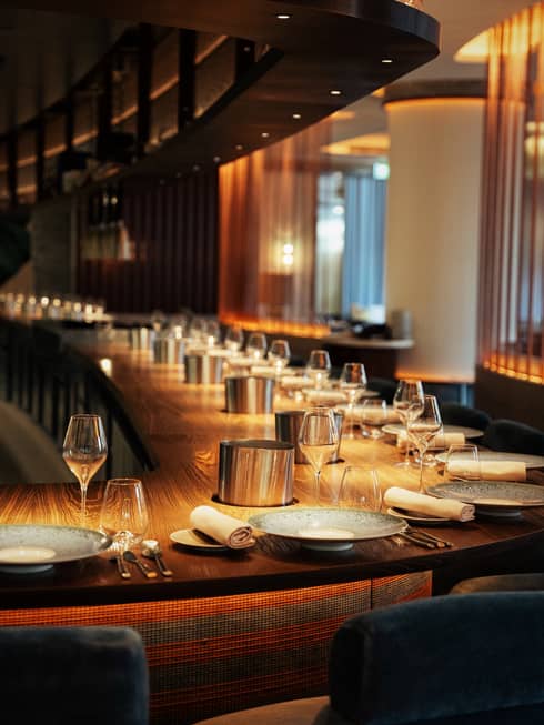 A long, curved solid wood bar with a dozen place settings in warm, golden lighting.