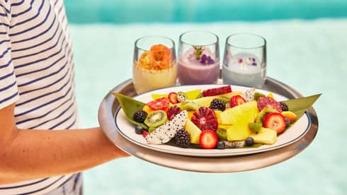 Person carrying tray of smoothies and fruit by pool