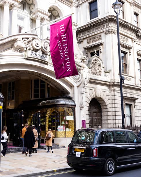 London-style black cab parked in front of a high-end pastry shop in a building with a Burlington Arcade sign and flag.