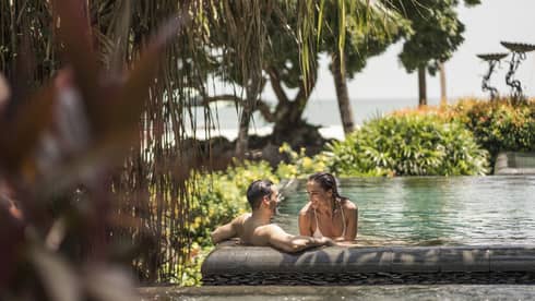 Smiling couple wades in outdoor swimming pool surrounded by tropical gardens