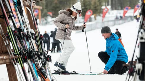 Hotel staff holds skis as woman holding poles steps in by ski rack, snowy hill