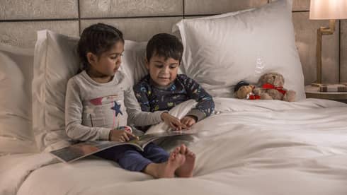 Young girl reads to her brother at bedtime, both in pajamas, a teddy bear propped on the pillow next to them
