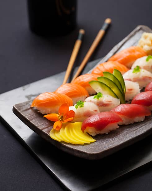 A wooden dish is topped with various colorful sushi, garnished with an orange flower