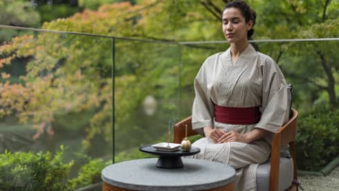 A woman sits on glass-enclosed balcony in lush setting with traditional Japanese robe on, meditating with an orin bell