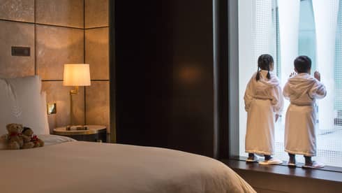 Young boy and girl in bath robes and slippers peer out the window at the city below, two teddy bears on the bed behind them