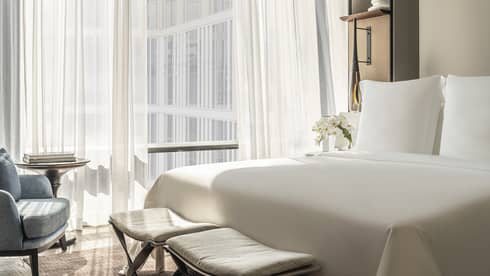 Hotel room with white bed and two benches at foot, facing blue chair, off-white wall by curtained window