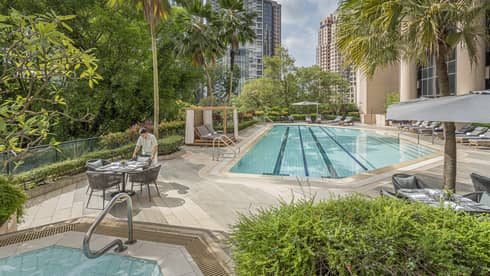 Outdoor pool area of a hotel, surrounded by lush greenery and tall buildings, with a staff member setting up a table beside a jetted tub and chaise lounges