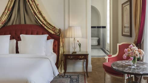 Hotel suite bedroom with bed furnished with white linens, red tufted headboard and curtains draped from the ceiling
