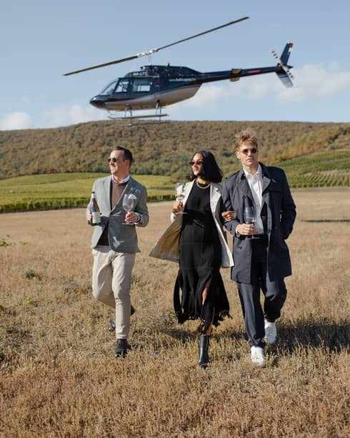 Three friends in chic outfits holding wine glasses walking in field with helicopter overhead