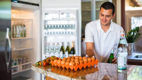 Smiling man holds green bottle in front of open fridge, fresh eggs on counter